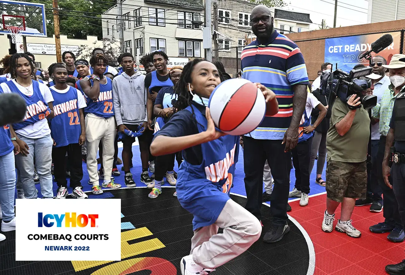 Shaquille O'Neal and a group of enthusiastic young basketball enthusiasts eagerly observe a young kid preparing to make a basketball throw. The newly renovated basketball court in Newark proudly features the Icy Hot logo on its floor. The lower-left corner of the image features a label displaying the Icy Hot Comebaq Courts logo, along with the event's year (2022) and location (Newark).