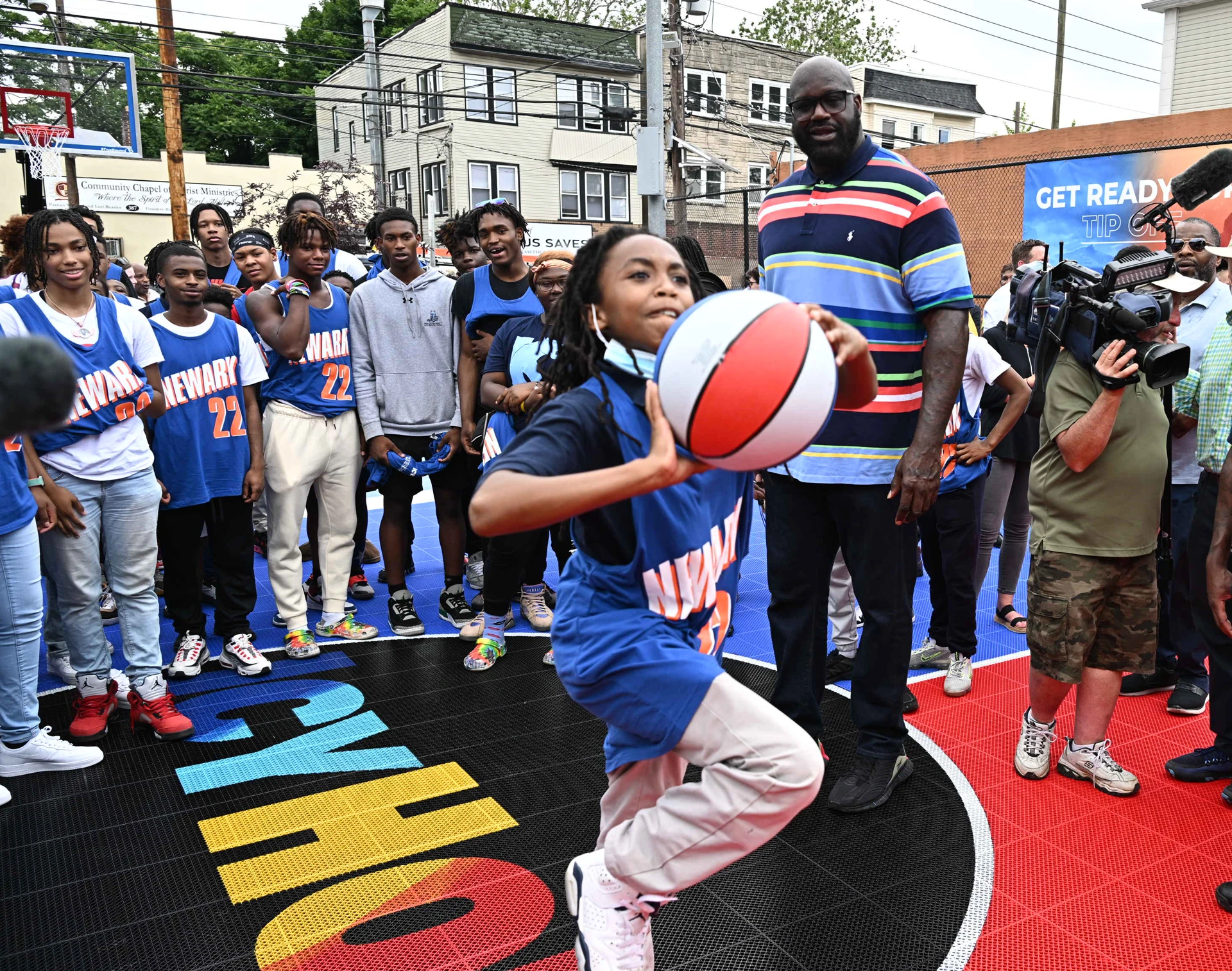 A moment from one of the Comebaq Courts' events. A young kid is playing basketball as Shaquille O'Neal and other kids look at her with attention.
