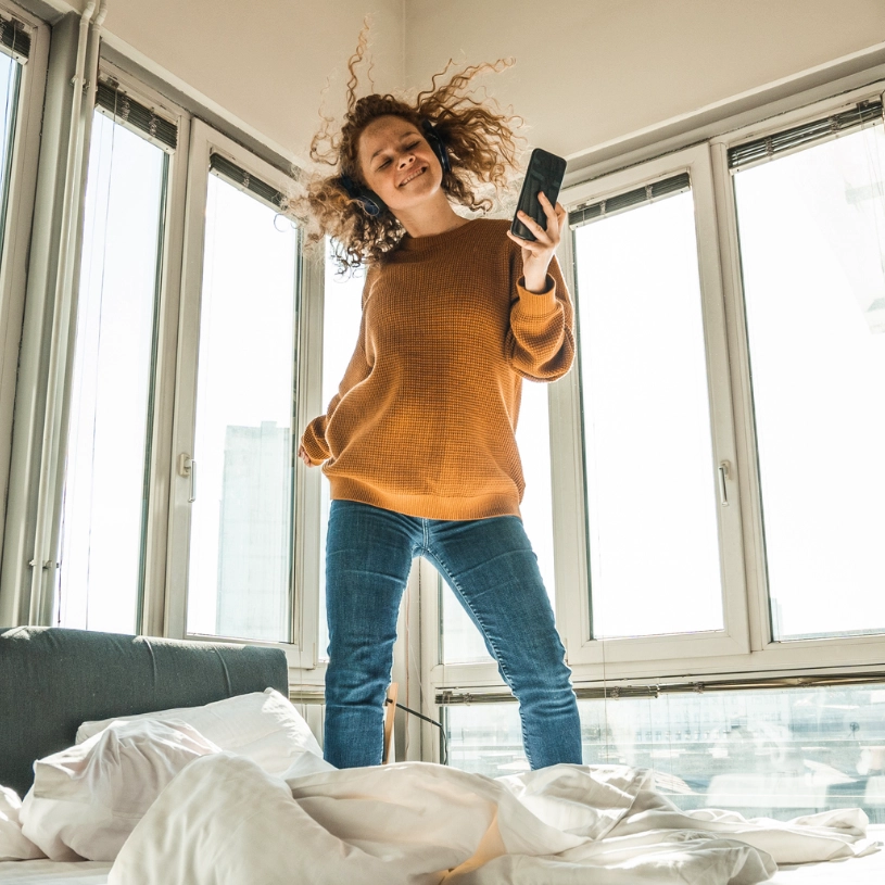 Glückliche Frau hört Musik mit Kopfhörern und tanzt auf dem Bett.