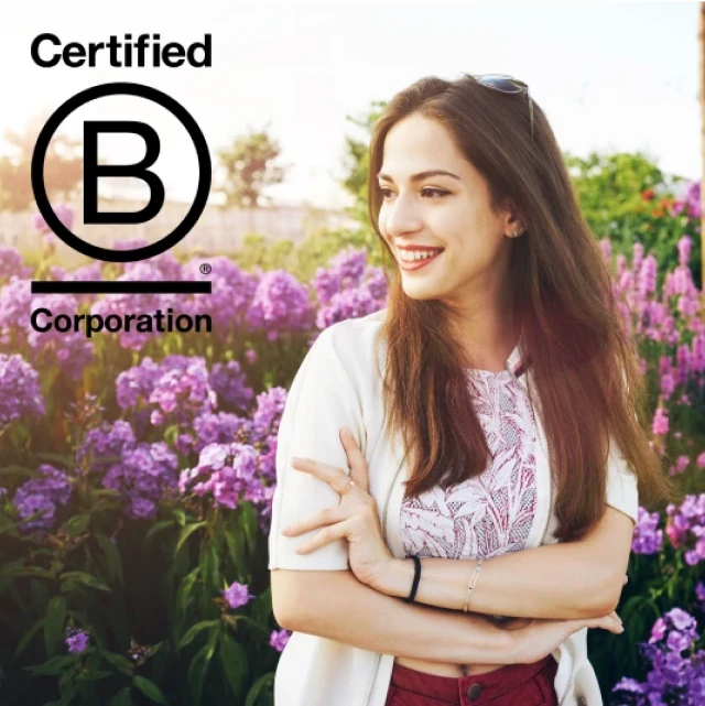 Eine junge Frau steht lächelnd vor einem Blumenfeld. Das Bild enthält das Logo der Certified B Corporation.
