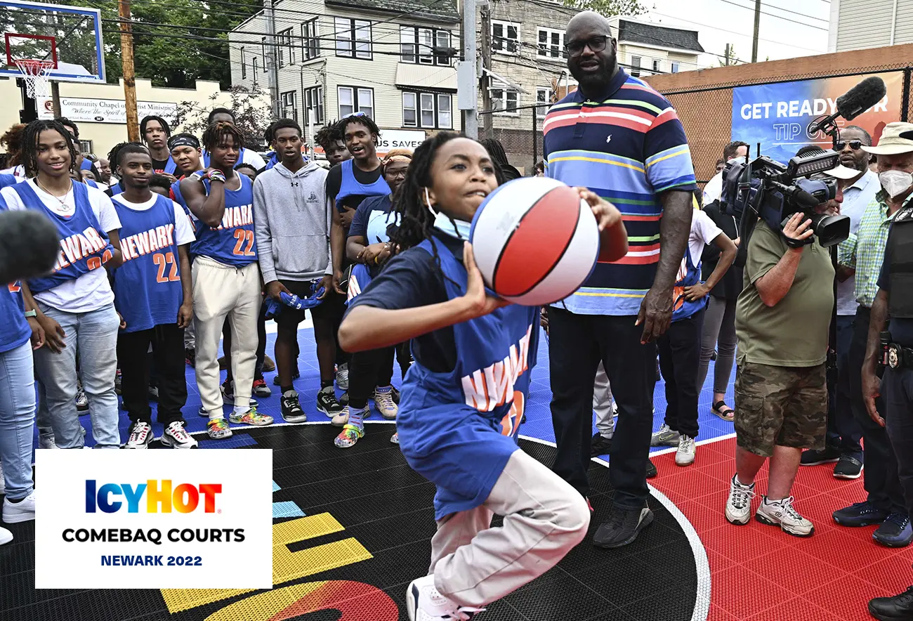 Shaquille O'Neal and a group of enthusiastic young basketball enthusiasts eagerly observe a young kid preparing to make a basketball throw. The newly renovated basketball court in Newark proudly features the Icy Hot logo on its floor. The lower-left corner of the image features a label displaying the Icy Hot Comebaq Courts logo, along with the event's year (2022) and location (Newark).