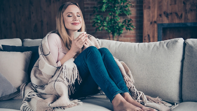A woman sitting cozily on a couch enjoying a drink out of a mug.